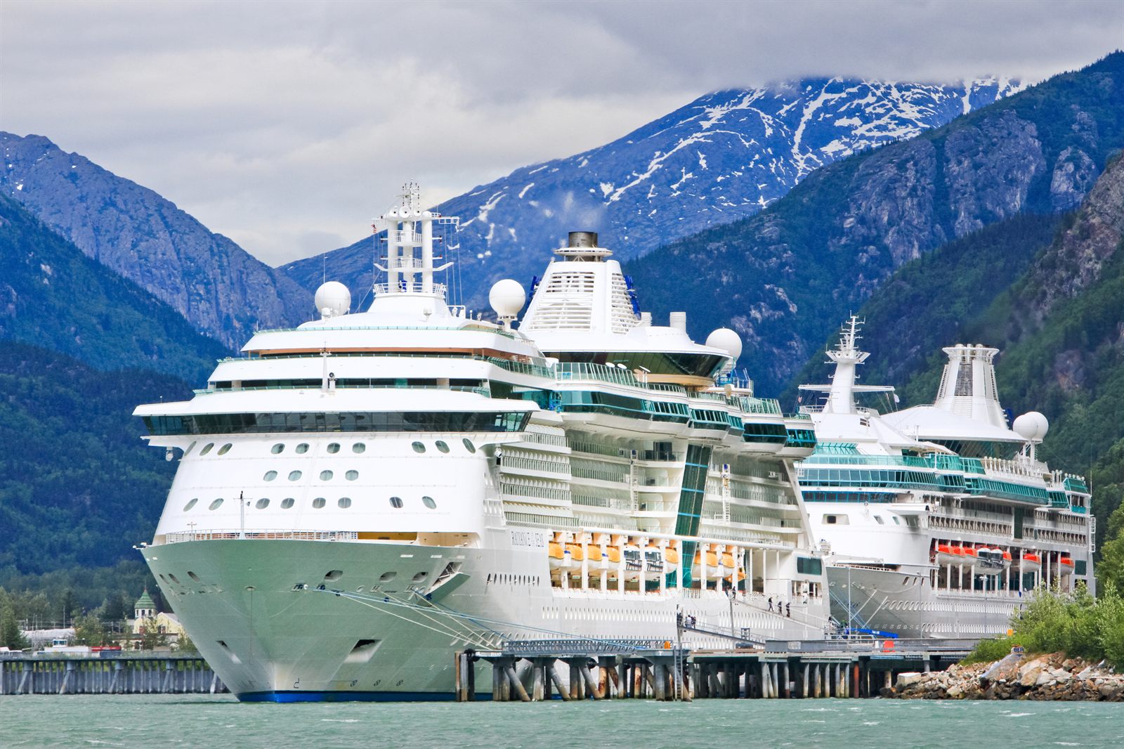 Two cruise ships docked below snowy mountains in an Alaska port